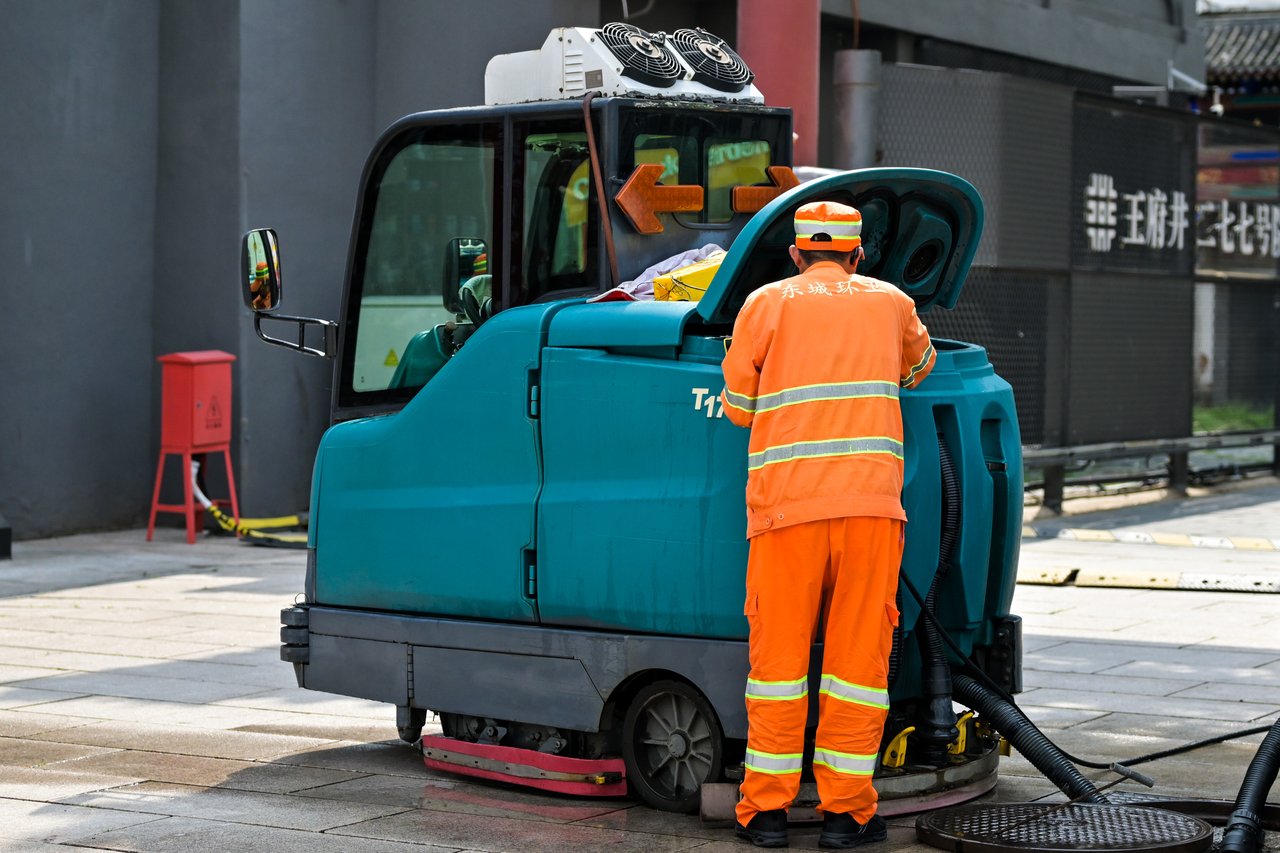 services-02 A street cleaner in uniform operates a cleaning machine on an urban sidewalk.