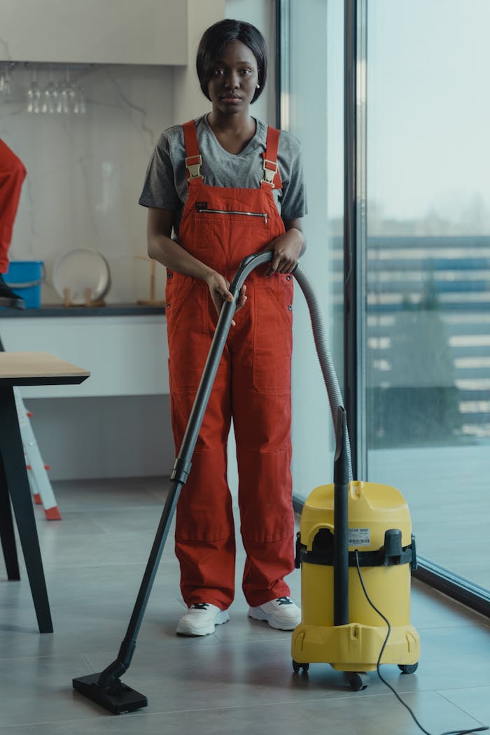 about-02 An adult woman in red coveralls operates a vacuum cleaner in a modern indoor setting, focusing on cleaning tasks.