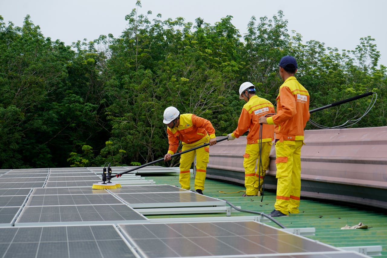hero-img-01 Three technicians in uniforms and hardhats clean rooftop solar panels in a renewable energy project.