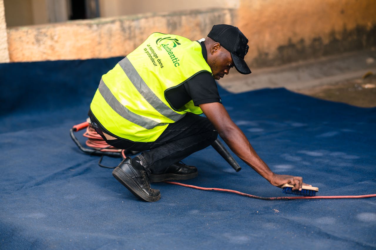 hero-img-02 A worker dressed in a safety vest cleans a blue carpet outdoors with a brush.